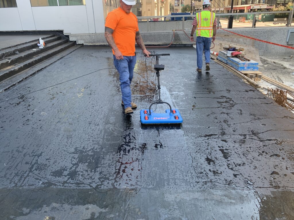 Technician moving an electronic leak detection scanning platform across the wet membrane on the deck at 100 North Broadway.