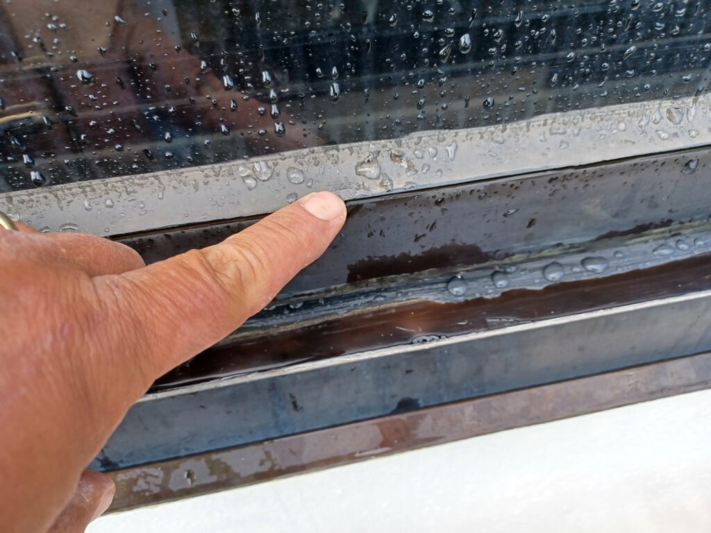 A hand points to standing water collected along the interior sill of a window frame during testing.