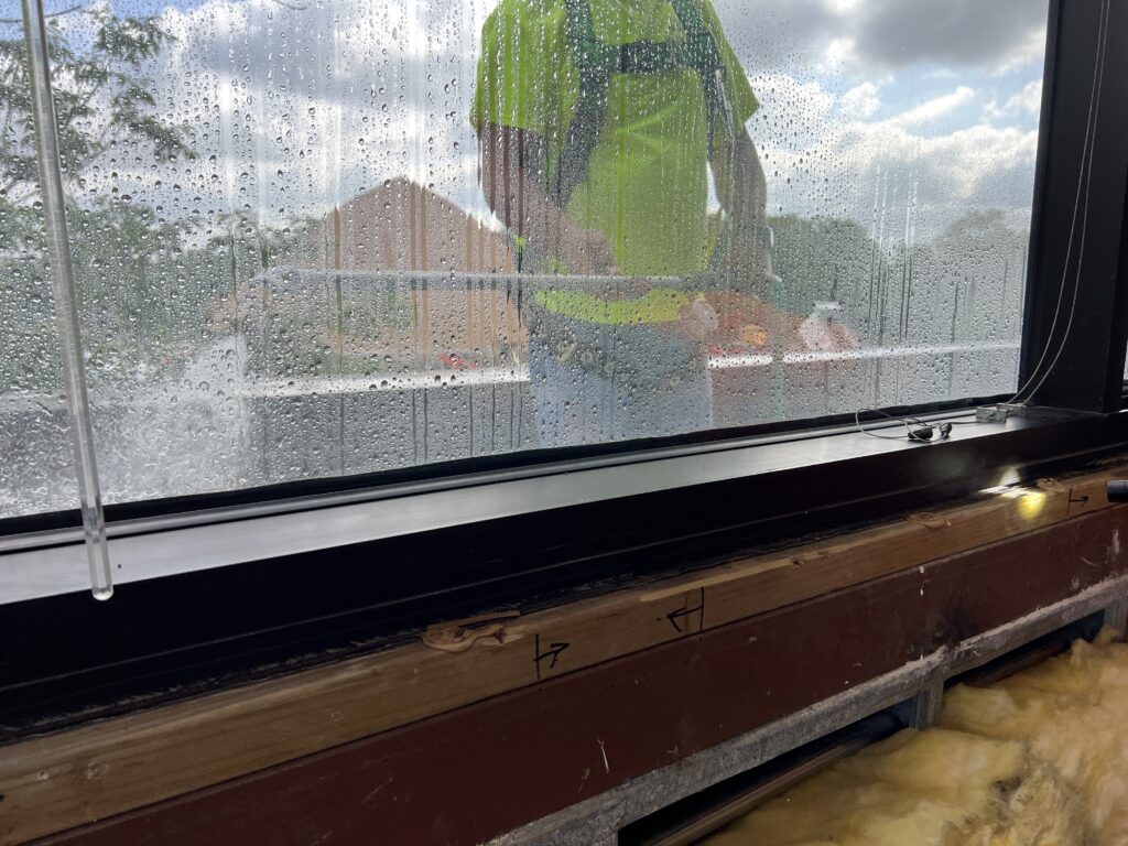 Water beading on the interior face of a window with a testing team member visible outside through the glass.