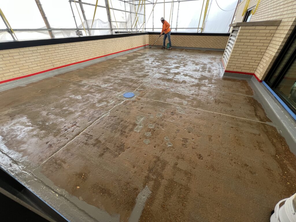 A worker applying water across a bricked-in terrace deck, membrane exposed, drain visible.