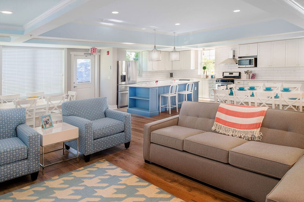 This open-concept residencial home for boys and girls features a neutral gray fabric sofa accented with a coral-and-white striped throw blanket, complemented by matching blue patterned armchairs and an adjacent kitchen with a bright blue island.