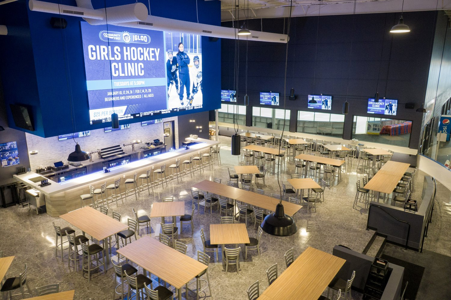 Eating area inside the recreation area. Plain wooden tables with stools facing a large tV screen