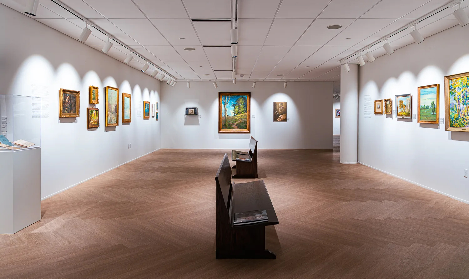 Gallery room in the Mercantile Library with framed landscape and portrait paintings under accent lighting, and wooden benches at the center.