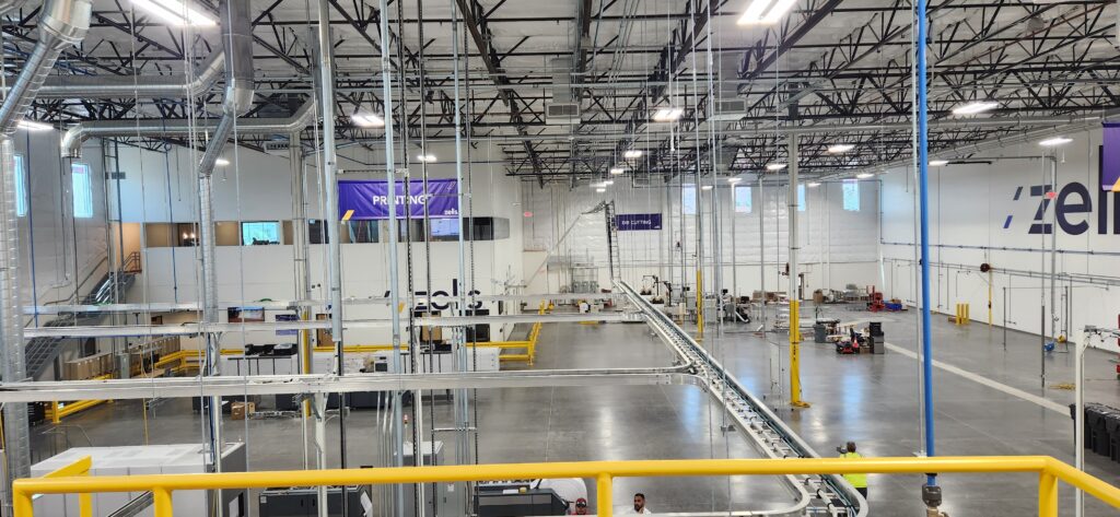 Interior of the Zelis print production floor showing the overhead air-driven conveyor, exposed ductwork, and lighting, with printing and die cutting zones.