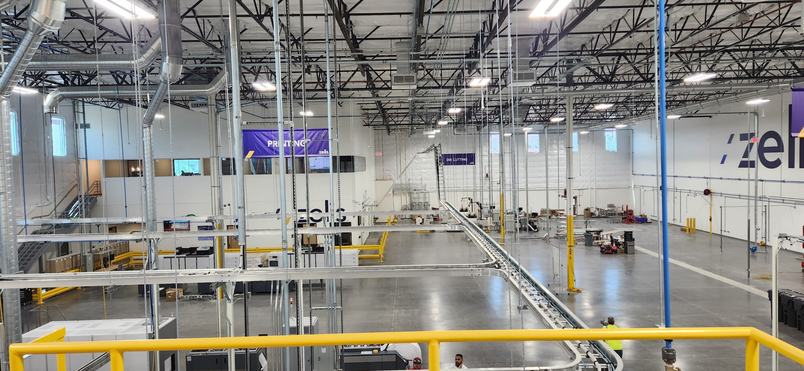 Interior of the Zelis print production floor showing the overhead air-driven conveyor, exposed ductwork, and lighting, with printing and die cutting zones.