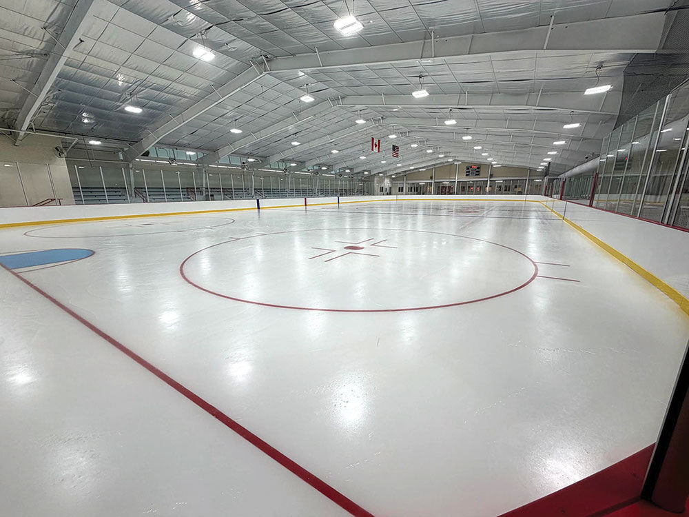 Freshly resurfaced ice at the renovated Kirkwood Community Center rink, with new dasher boards and glass, red and blue hockey markings, LED lighting overhead, and bleachers along the left side.