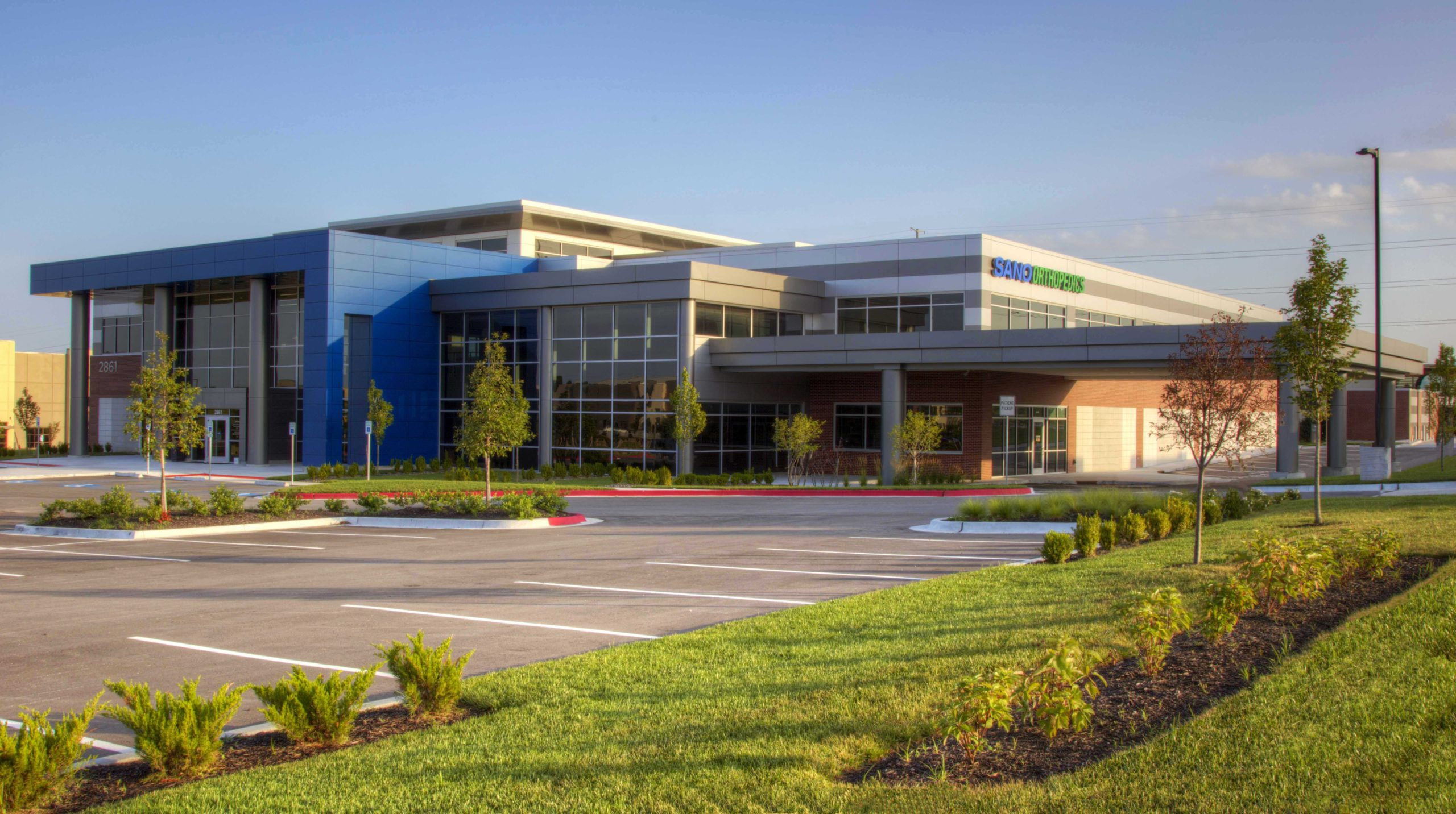 Two-story surgery center and medical office building in Lee’s Summit, Missouri, with blue metal panel, glass curtain wall, and a covered patient drop-off along the front parking lot.
