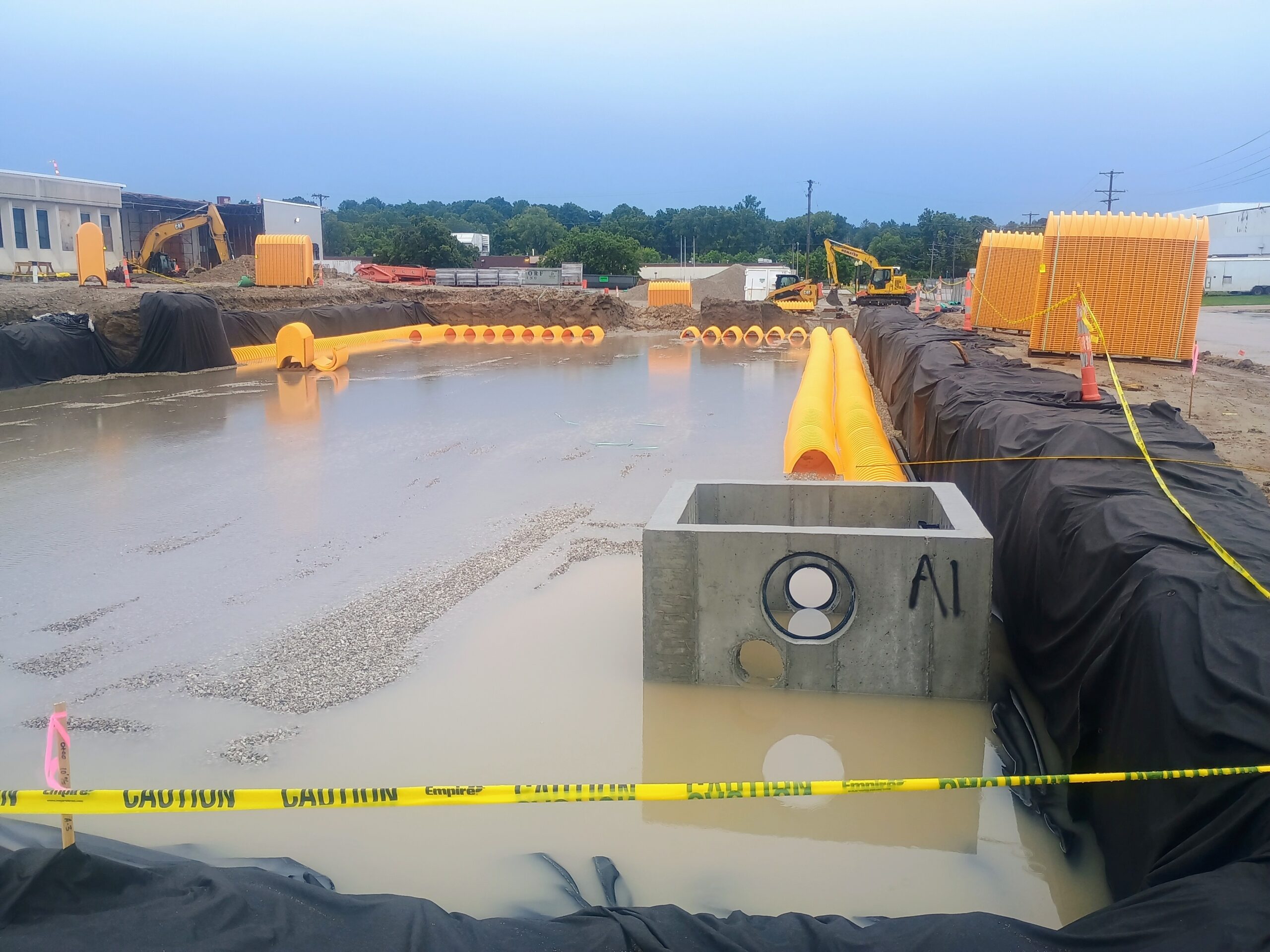 Excavated stormwater detention basin holding rainwater, with a concrete inlet structure and orange chamber rows at the Enterprise loading dock site.