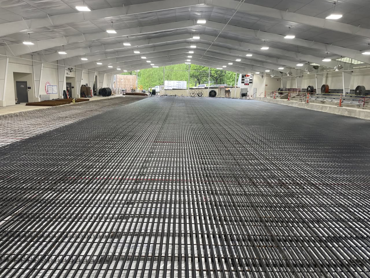 Refrigeration piping laid in a grid across the Kirkwood ice rink floor before the concrete slab pour, with construction equipment at the far end.