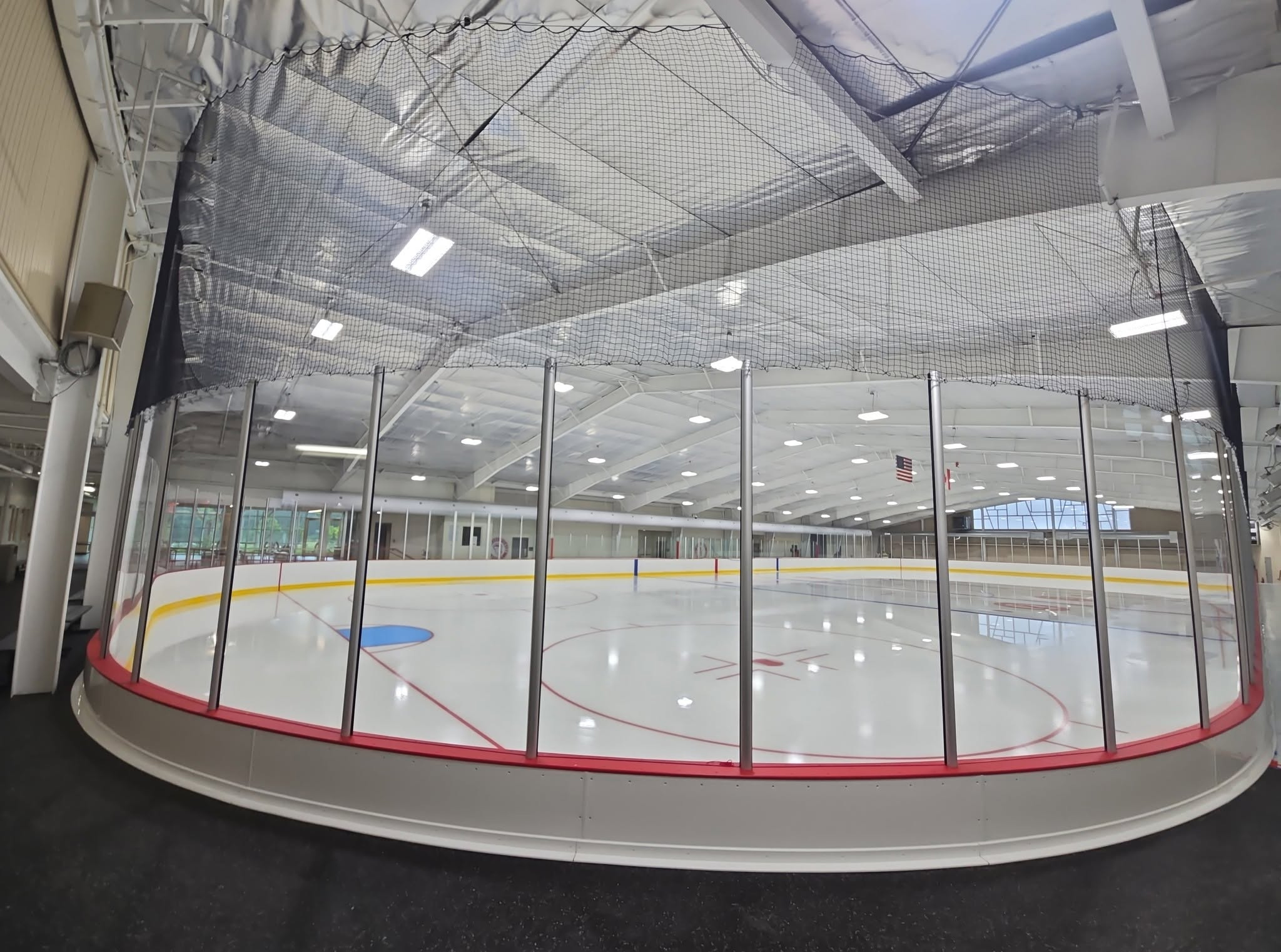 Wide view of the renovated Kirkwood ice rink through the new dasher boards and glass, with protective netting overhead and fresh ice markings.