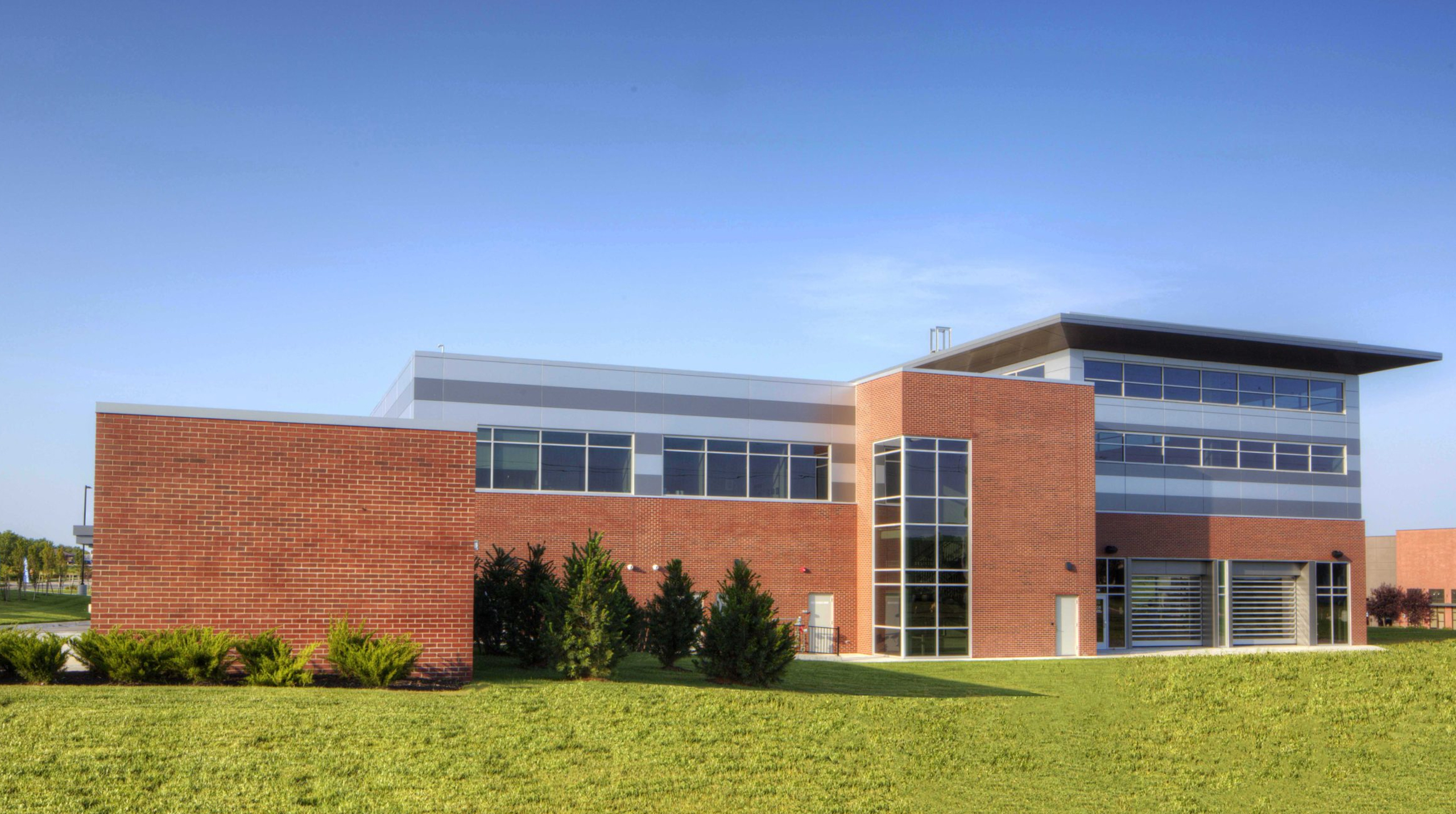 Side view of the Lee’s Summit surgery center showing red brick and metal panel walls, a glass stair tower, and overhead coiling doors at the physical therapy area.