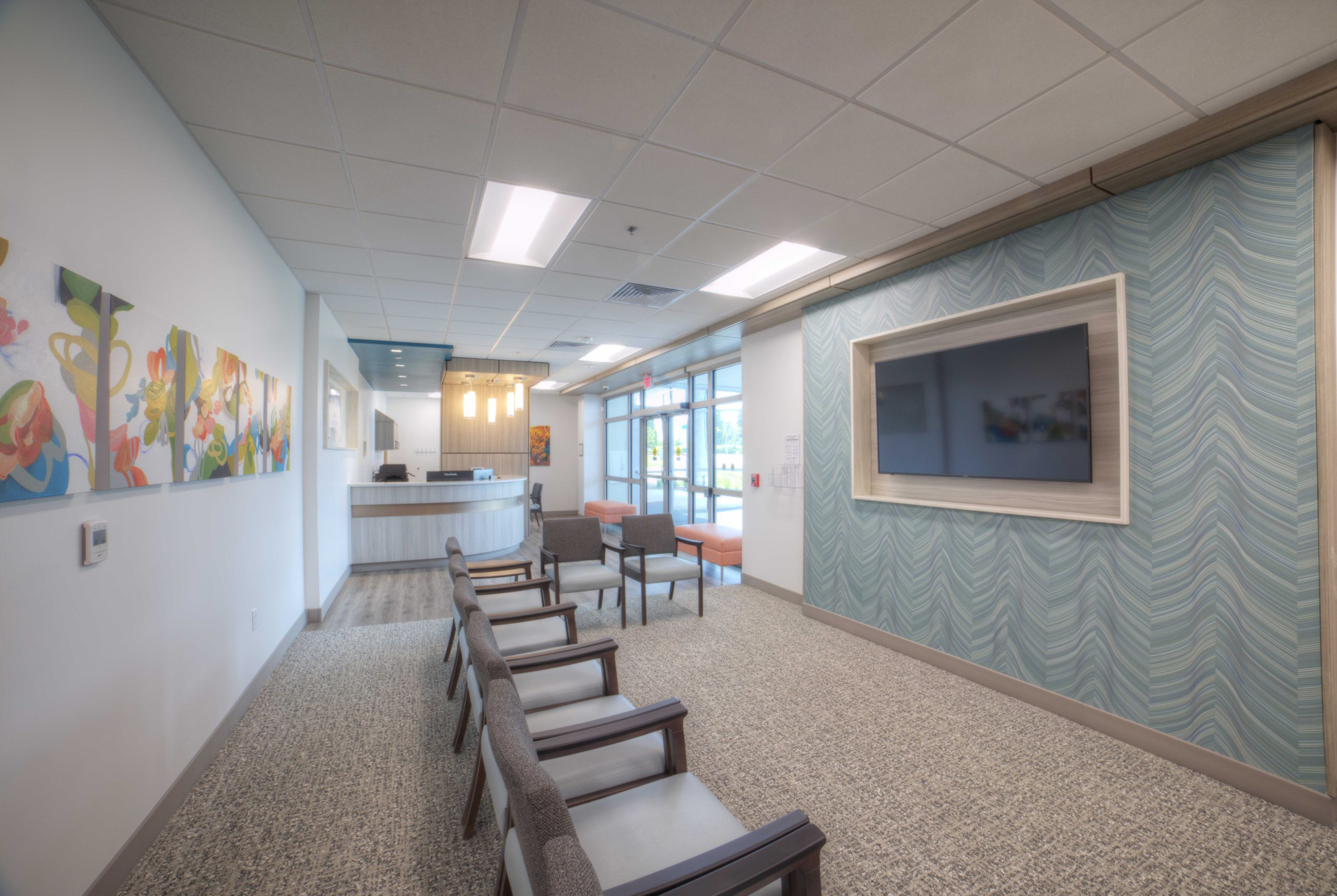 Patient waiting area with upholstered chairs, a textured blue accent wall with a mounted display, and a reception desk near the glass entry.