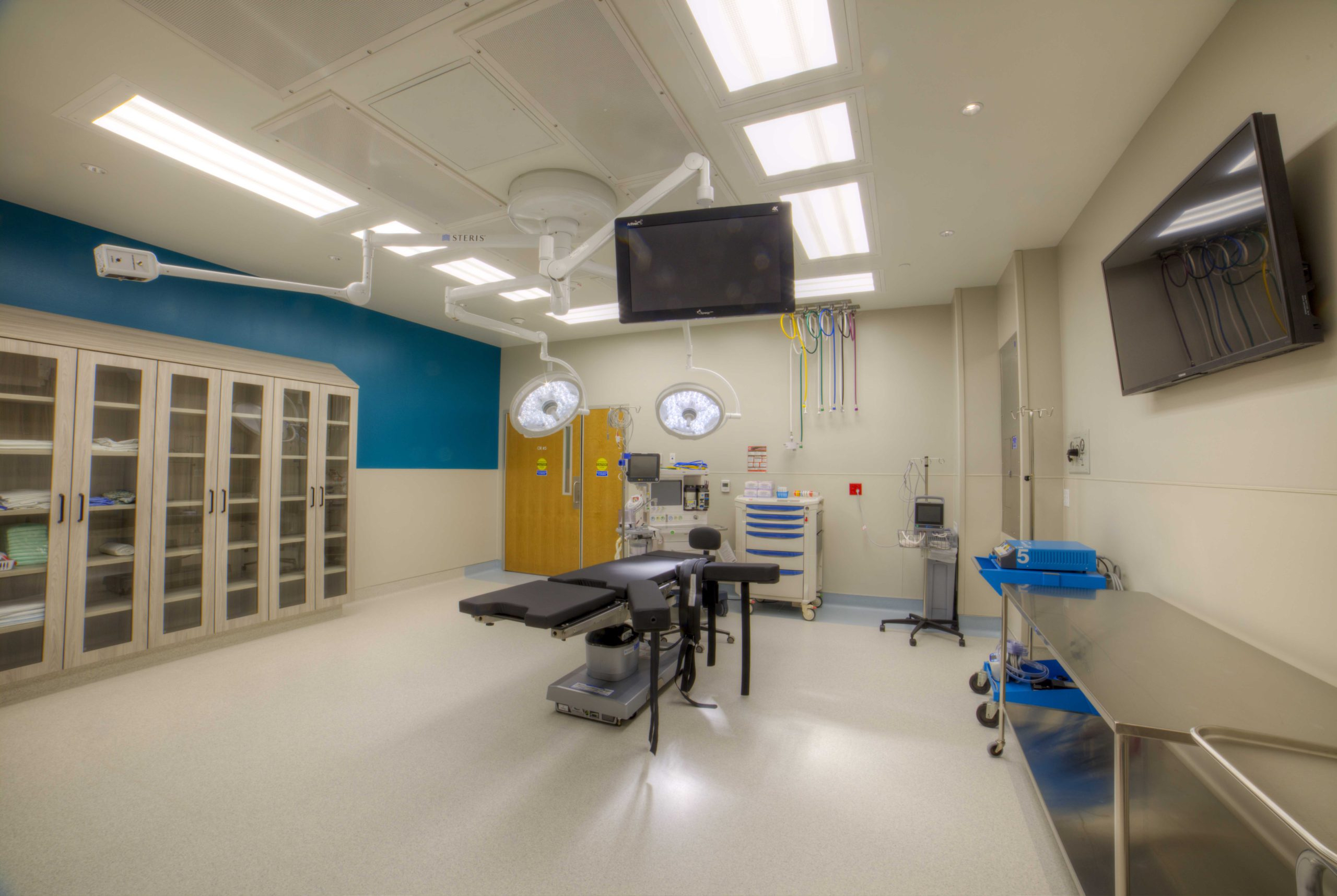 Operating room with surgical lights, ceiling-mounted equipment booms and monitors, an operating table, and built-in glass-front supply cabinets.